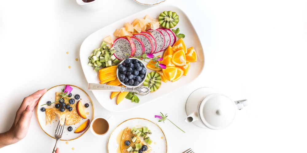 A person having a healthy breakfast consisting of crepes with blueberries and slices of peach, and a plate with an assortment of fruits like dragon fruit, kiwi, orange, and blueberries, accompanied by a pot of tea and a cup.
