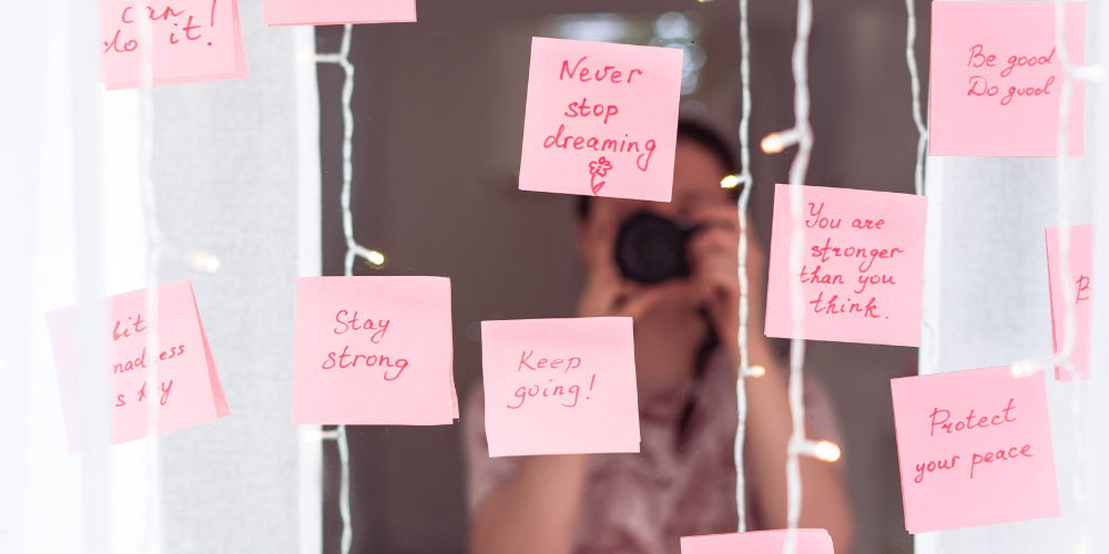 An image featuring a collection of inspirational sticky notes on a mirror with a person taking a photograph in the reflection. The notes contain positive affirmations 