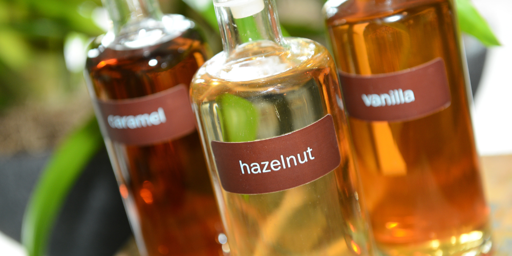 Three glass bottles of coffee syrup labeled "caramel," "hazelnut," and "vanilla," with the hazelnut one in focus in the center. The bottles are set against a blurred natural background, possibly with green plants, suggesting an organic or fresh setting.