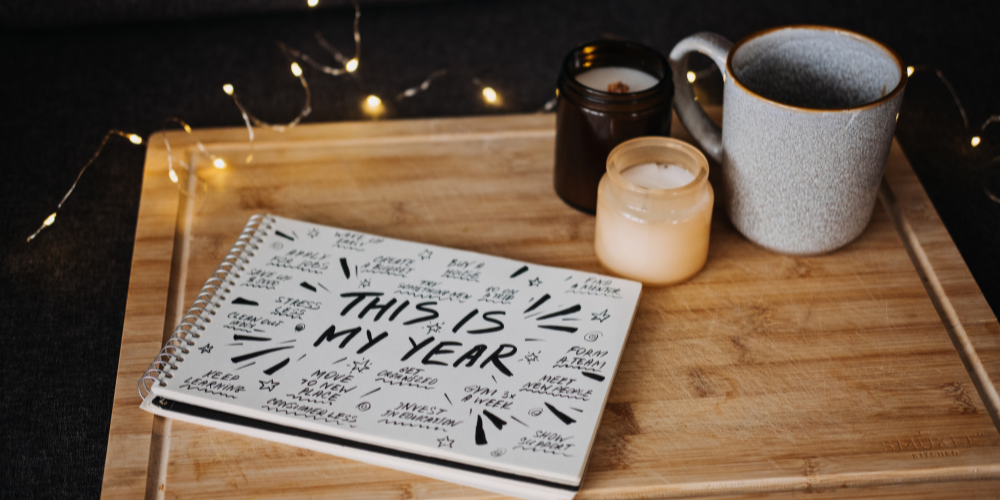 The image shows a notebook with "THIS IS MY YEAR" written on it, a mug, and a candle, all on a wooden surface, with fairy lights adding a warm ambiance.