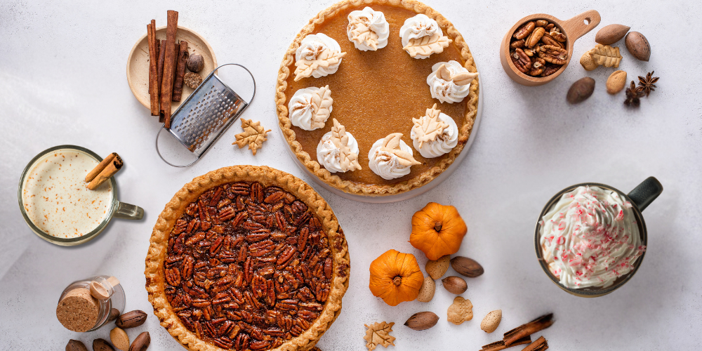 Top-down view of festive fall and winter holiday treats, including a pumpkin pie with whipped cream, a pecan pie, and mugs of eggnog and peppermint mocha. The image is surrounded by seasonal ingredients like cinnamon sticks, nuts, and small pumpkins.