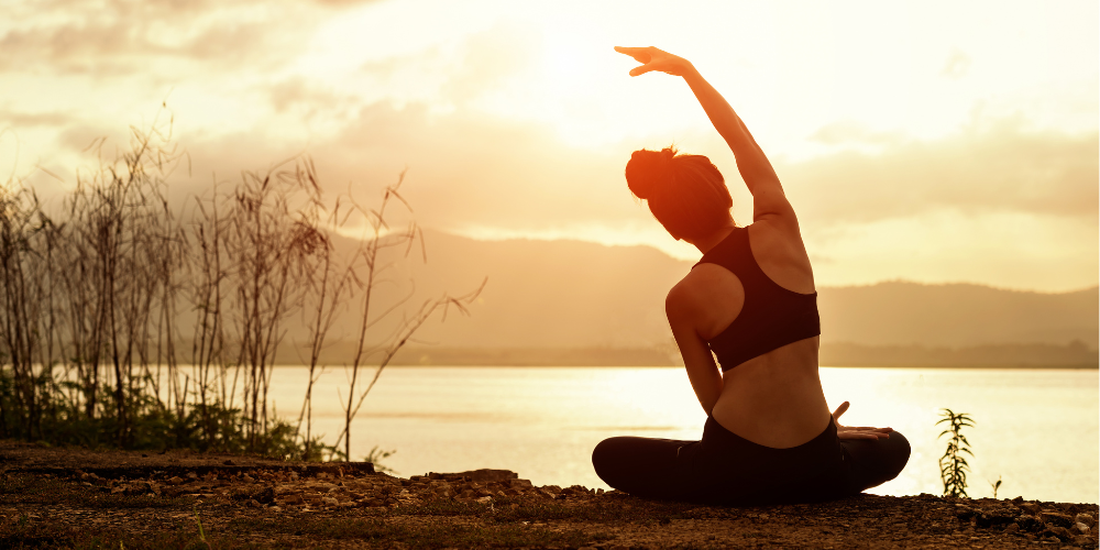 A woman practices yoga by a serene lakeside at sunset, stretching her arm towards the sky.
